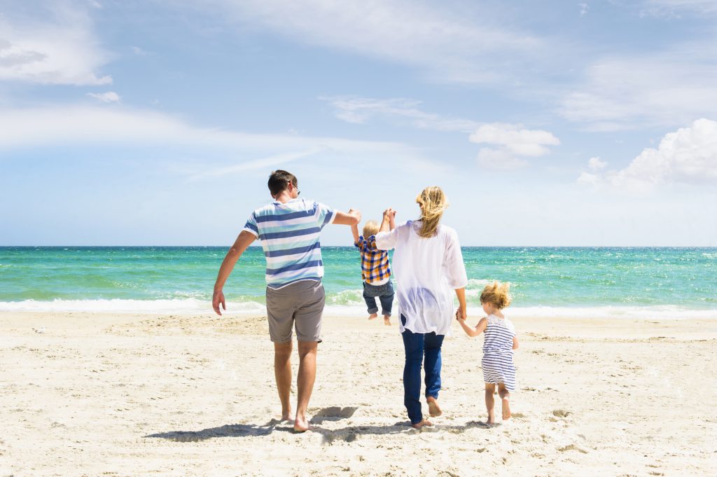 Caucasian family swinging son by arms at beach with crystal-clear water and white sand beach